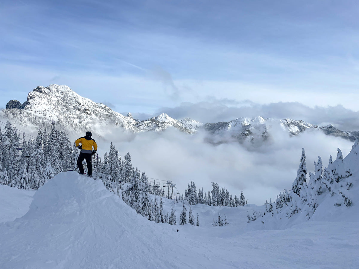 Person in yellow jacket standing on a snow-covered mountain with a scenic view of mountains and clouds.